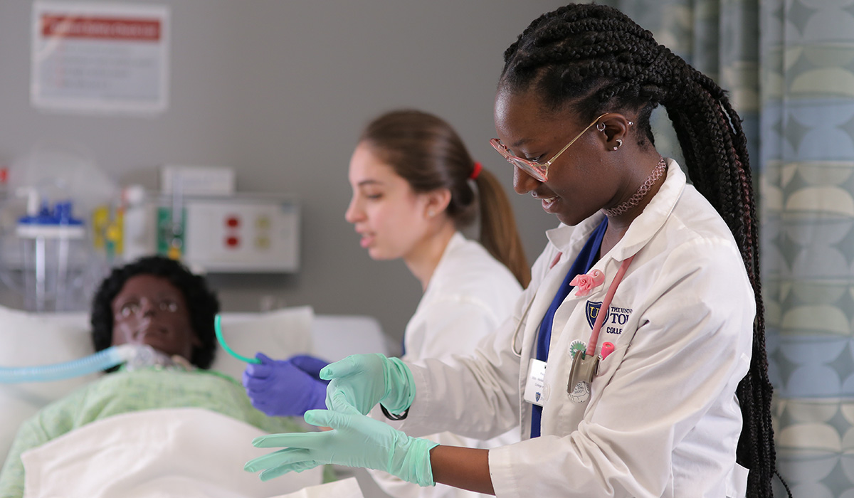 Two nursing students wearing medical clothing in a hospital room looking at a test doll in a hospital bed