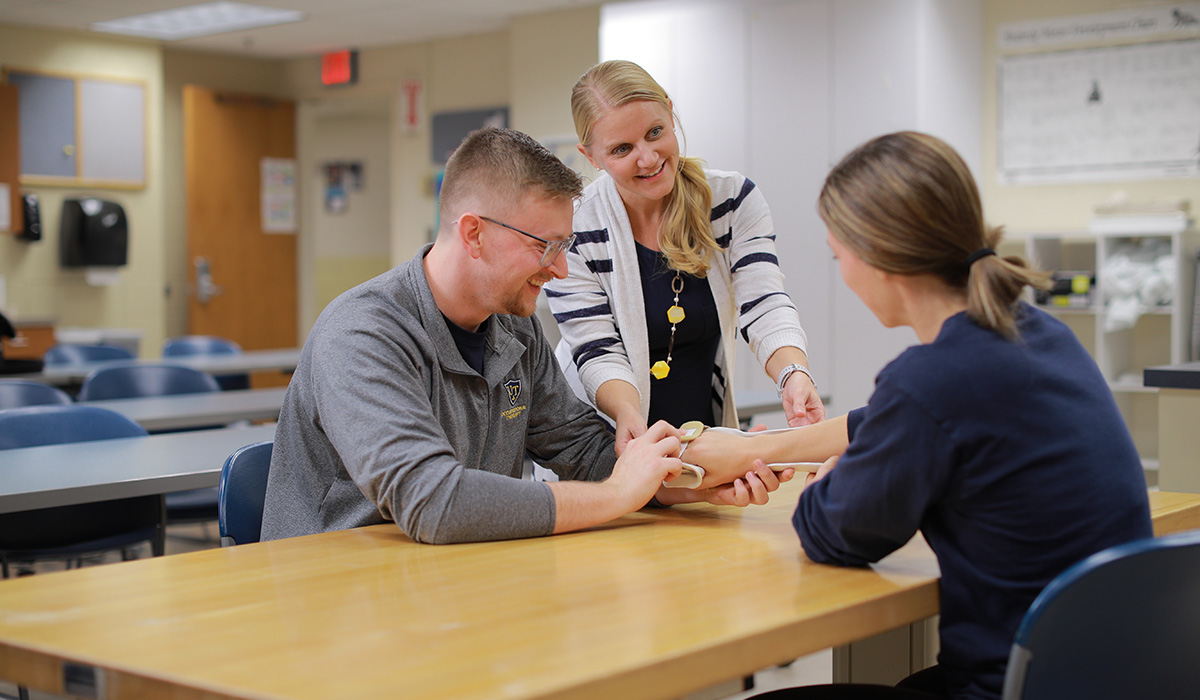 3 people sitting at a table in a classroom using medical equipment on the woman's arm