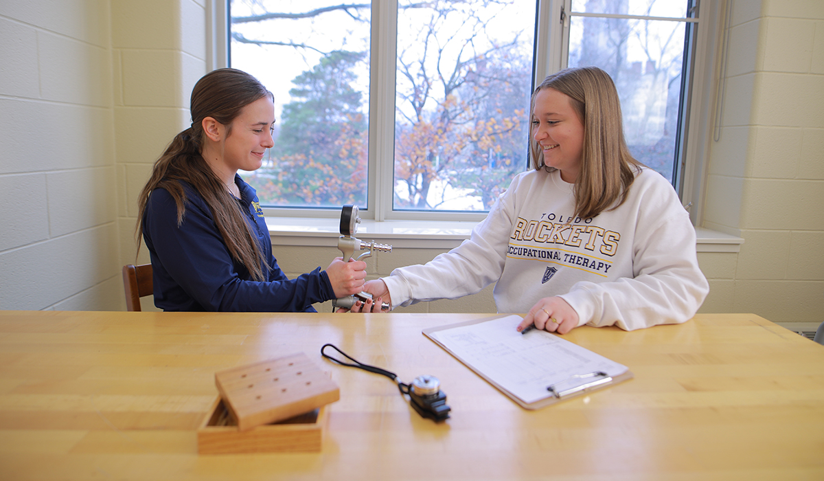 two otd students in a class room sitting at a table, both students are holding onto medical device