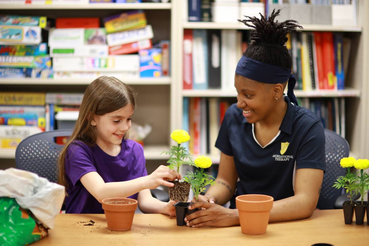 recreation therapy student and a child sit together at a table assembling a pot for a flower