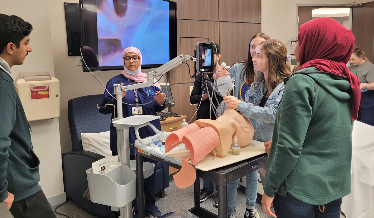 a group of respiratory care students and a faculty member in a medical classroom. One student is using medical tools on a practice mannequin while other students watch