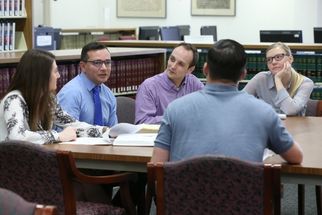 Five adults seated around a table in a library or study setting, engaged in discussion with open books in front of them. Bookshelves line the background.