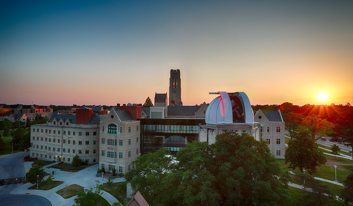 An image of UToledo's campus during the summer. There is a clear blue sky and the sun is setting in the background.