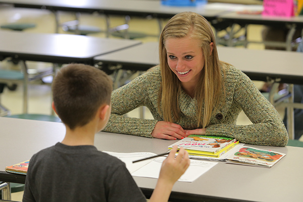 A woman and young boy sitting at a table. The young boy is holding a pencil working on a paper worksheet while the woman has children picture books in front of her.