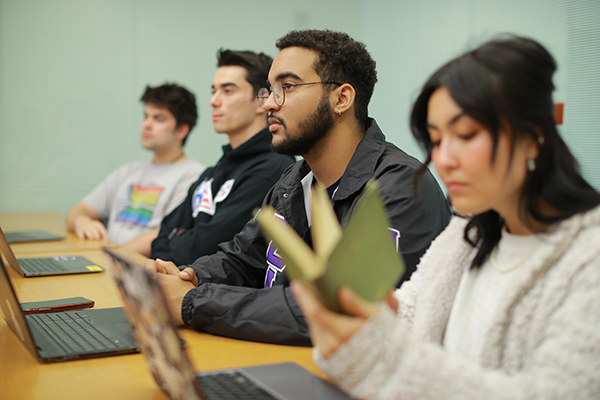 english students in a classroom sitting at a desk with laptops on the table and books in their hands