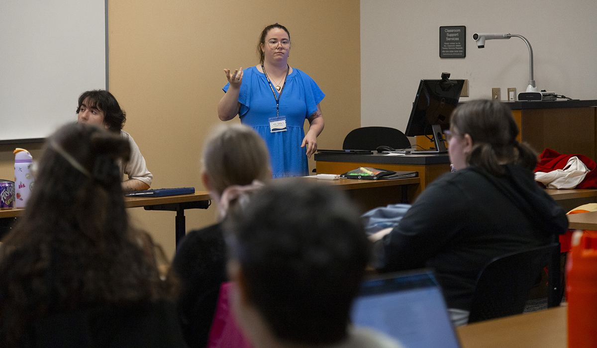 A woman in a blue dress standing in front of and talking to a classroom full of history stundents