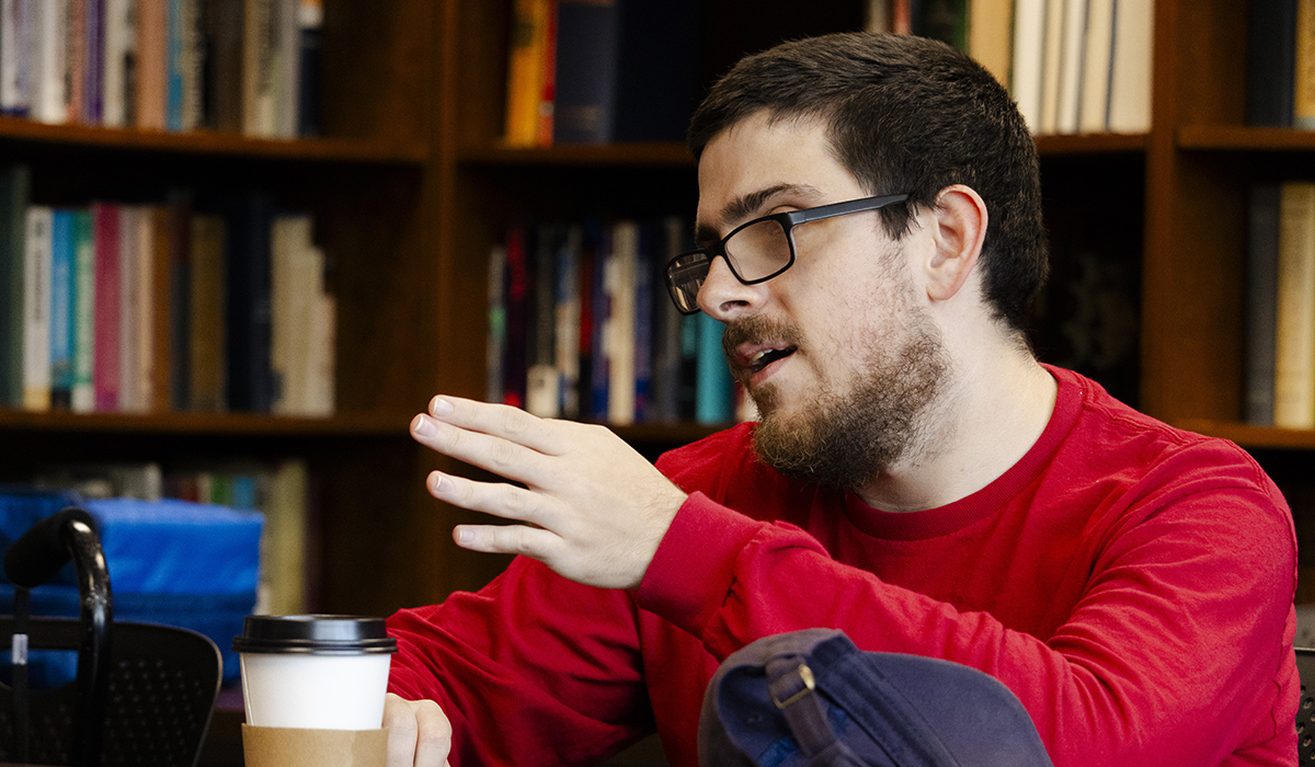 Student wearing a red shirt and glasses sits at a table in a library, gesturing while speaking during a discussion. A coffee cup, smartphone and backpack rest on the table, with bookshelves filled with books in the background.