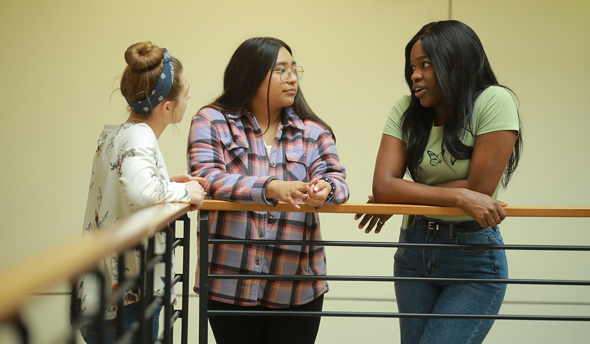 3 women and gender studies students leaning on a railing in a building talking to each other