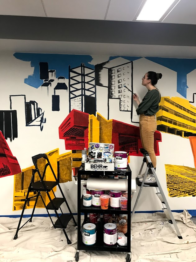 An Experiential Learning Artist painting a cityscape mural on the 2nd floor of Carlson Library.
