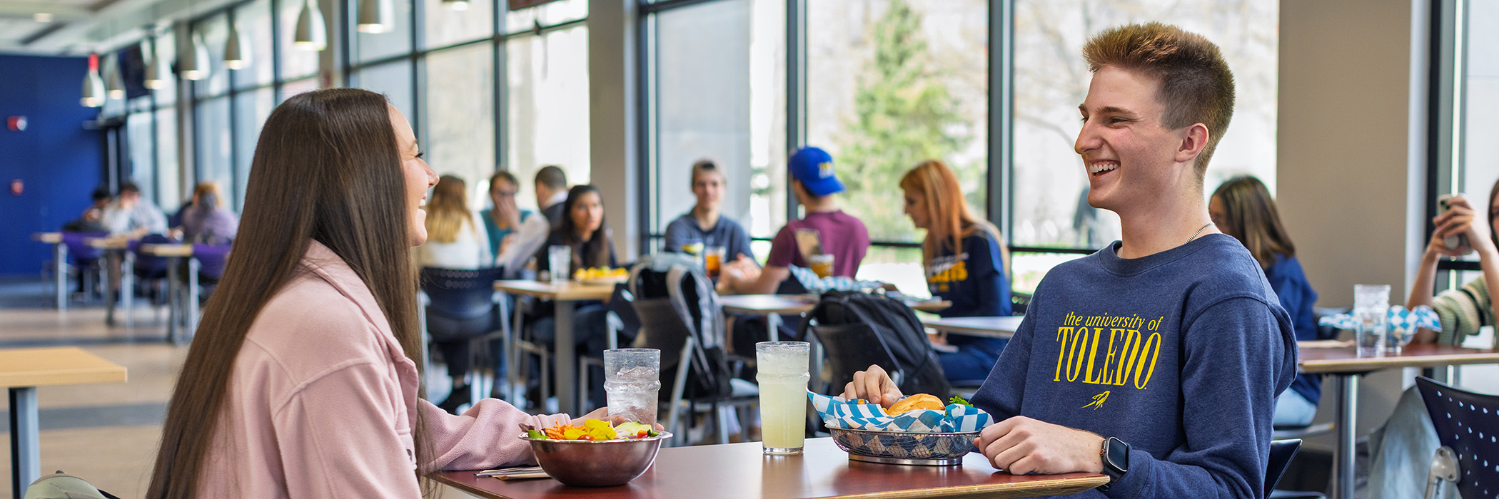 Two students share a meal and smile at a table in a bright dining hall.