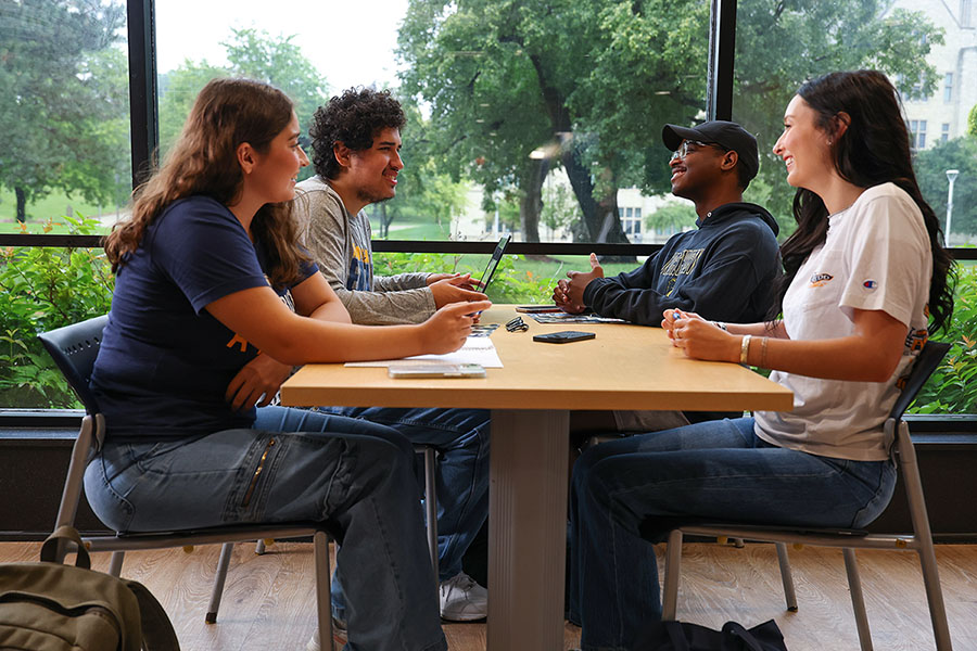 Four young adults talk around a table in the student union food court beside large windows overlooking centennial mall.