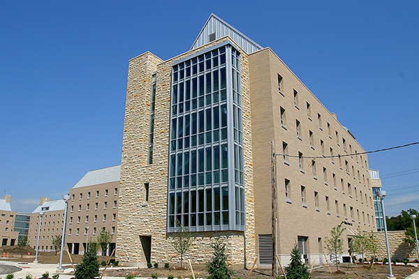 Multi-story residence hall with tan brick, stone walls, and a tall glass-fronted central section under a clear blue sky.