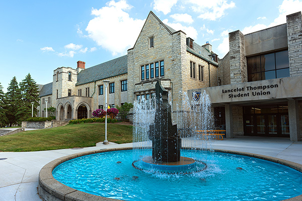 Lancelot Thompson Student Union behind the fountain, with trees and landscaping under a bright blue sky.