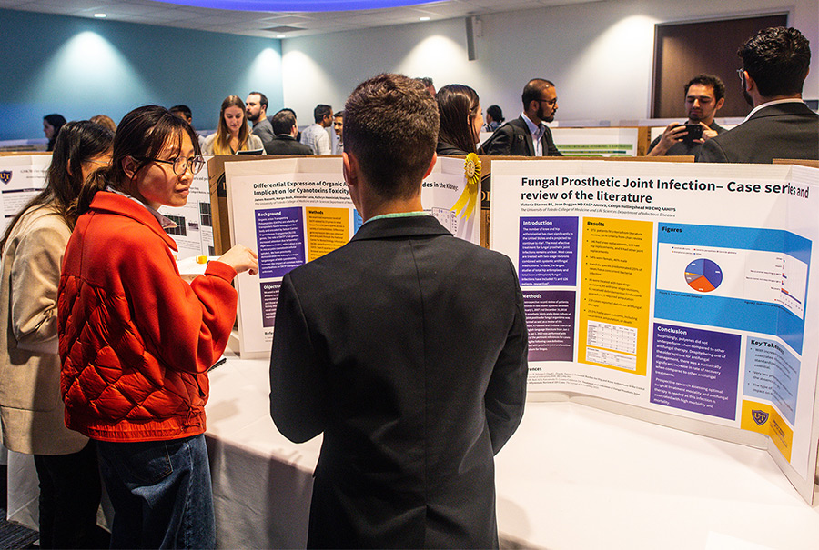 Students discuss their research posters at the 3rd Annual Research Symposium. Posters are pictured on tables inside the Simulation Center. 