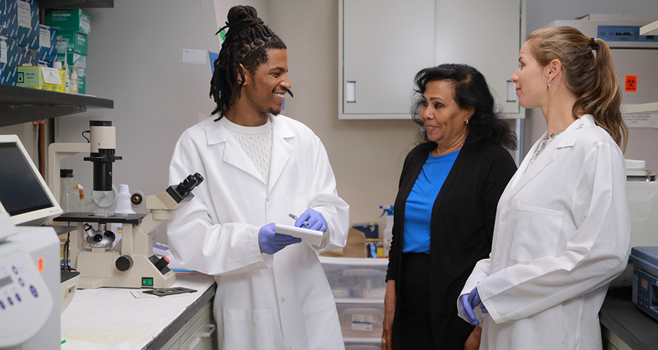 A faculty member speaks with two students wearing white coats in a lab setting