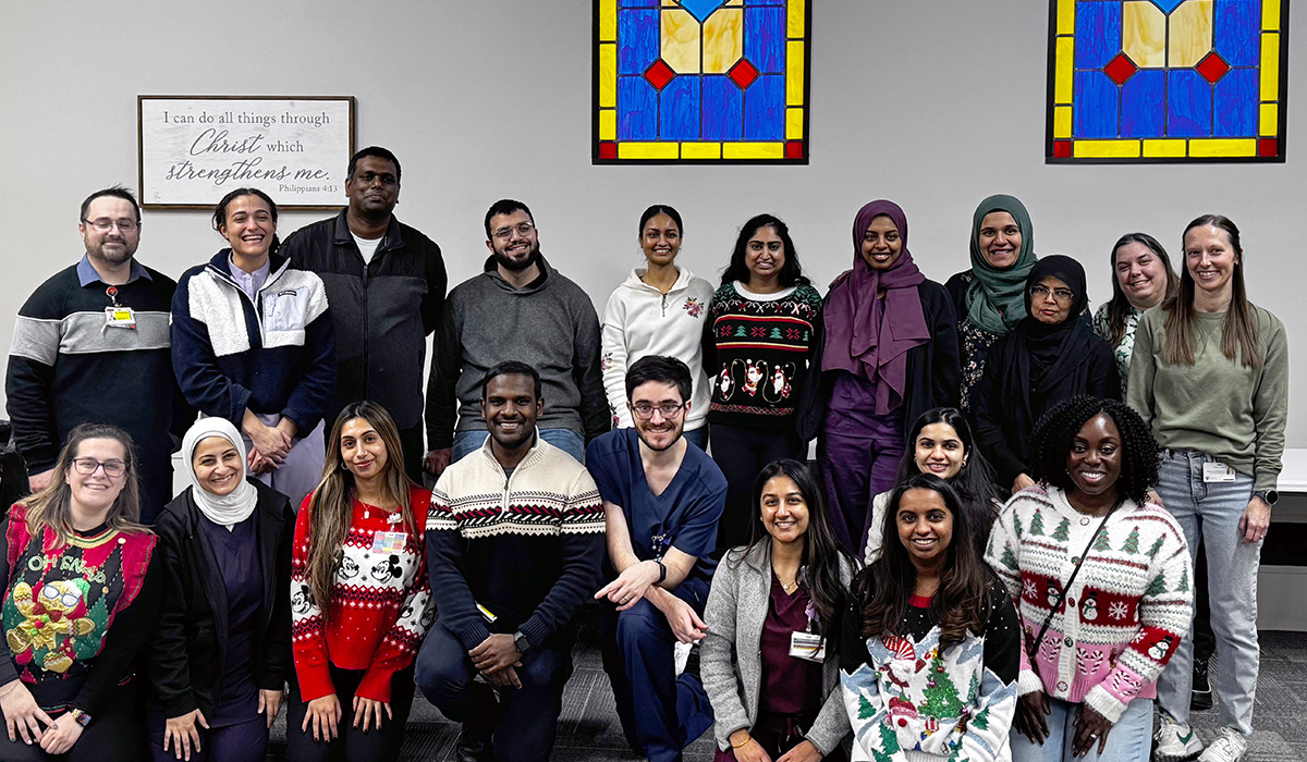 A large, diverse group of residents and staff pose together indoors, smiling in front of stained glass windows, many wearing festive holiday sweaters.