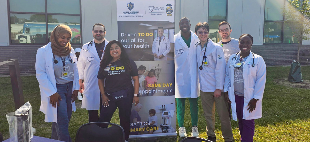 A group of medical residents and staff stand outdoors beside a University of Toledo Health display promoting pediatric primary care and same-day appointments during a community outreach event.