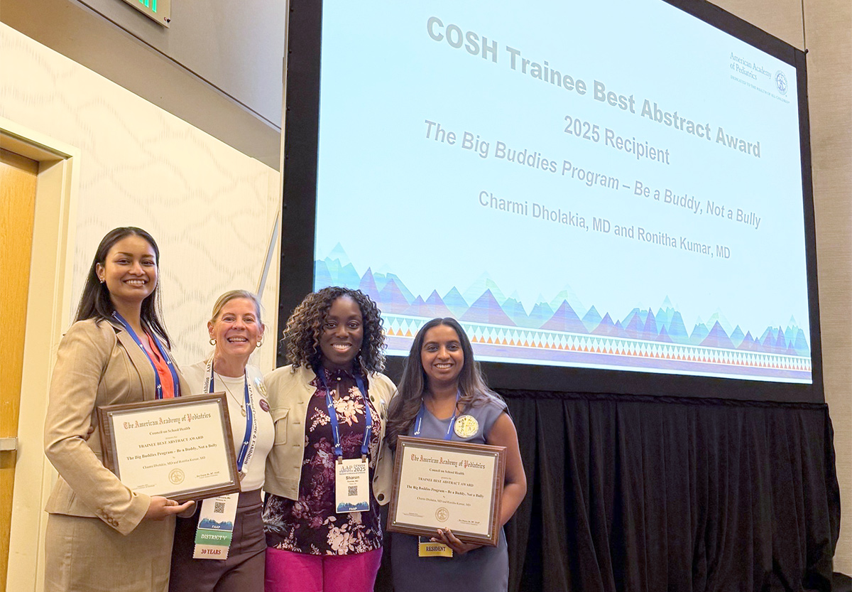 Four individuals stand onstage at a conference holding framed certificates, recognized as recipients of a trainee abstract award, with a presentation screen behind the