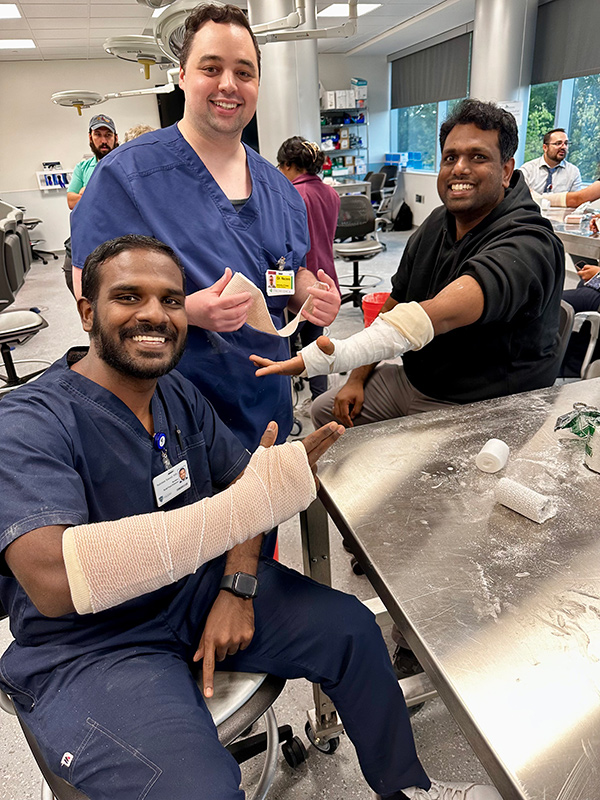 Three medical residents in a clinical skills lab smile while practicing arm casting, with one resident’s forearm wrapped in bandage material and supplies spread across a table.