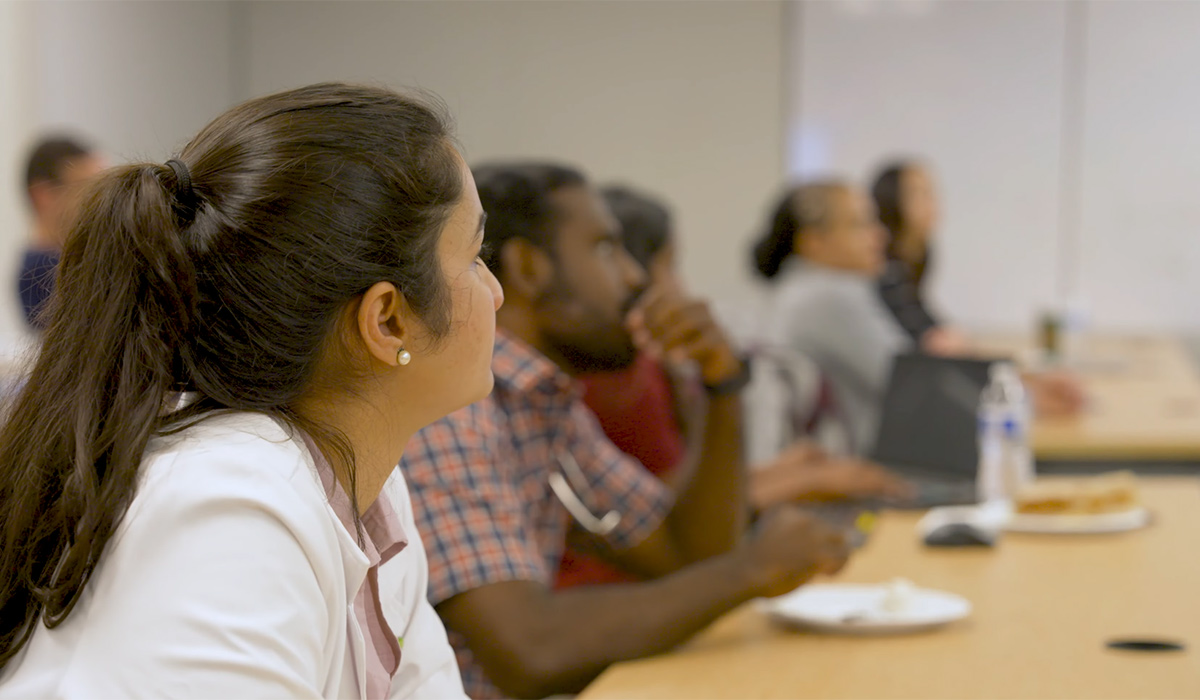 Medical residents sit at a conference table listening attentively during a classroom session, with laptops, notebooks and refreshments visible.