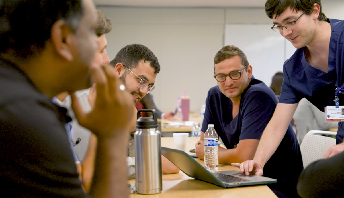 A group of residents collaborate around a laptop during a small-group session, discussing and reviewing information together.