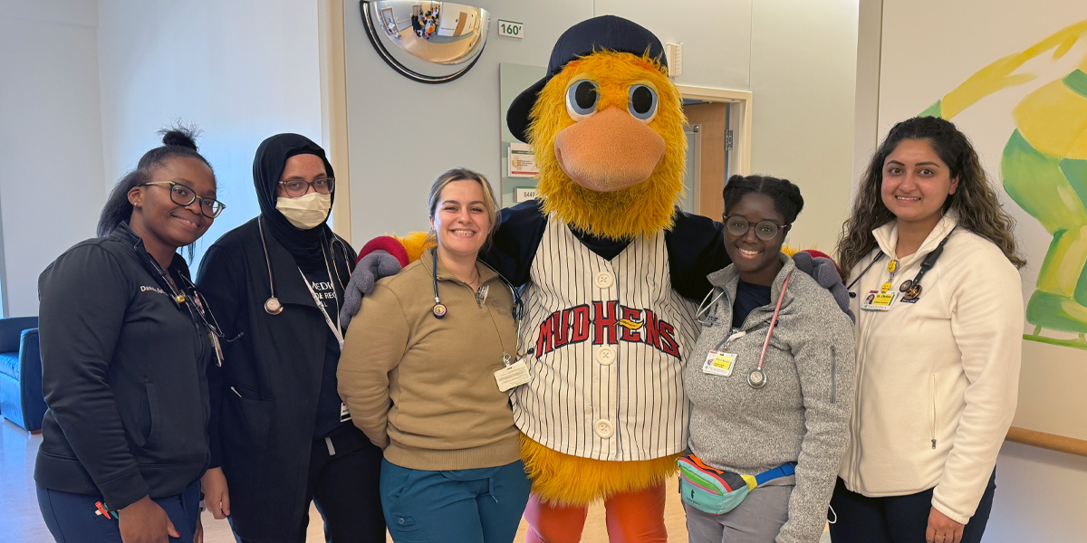 A group of pediatric residents pose in a hospital hallway with Muddy the Mud Hens mascot, smiling together for a photo.