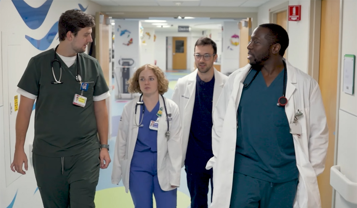 Four medical residents walk together down a brightly colored hospital hallway, wearing scrubs and white coats, engaged in conversation during rounds.