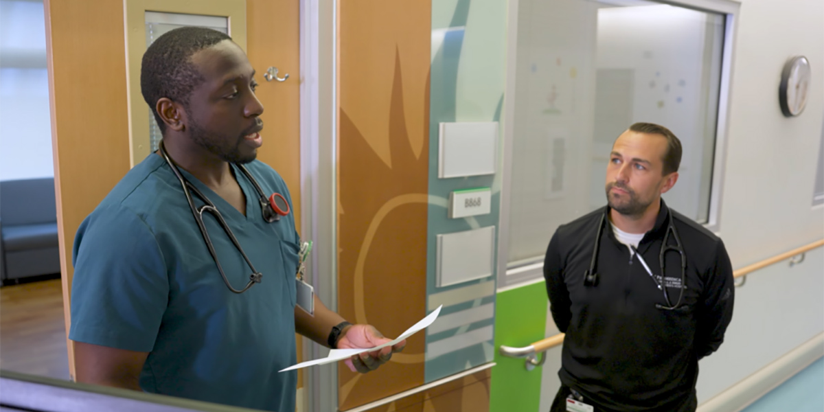 A pediatric resident in scrubs reviews a paper while speaking with another clinician in a hospital hallway.