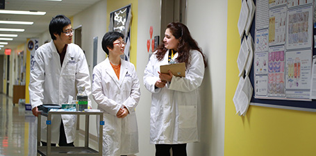 Three people in white coats hold a conversation while walking through a hallway.