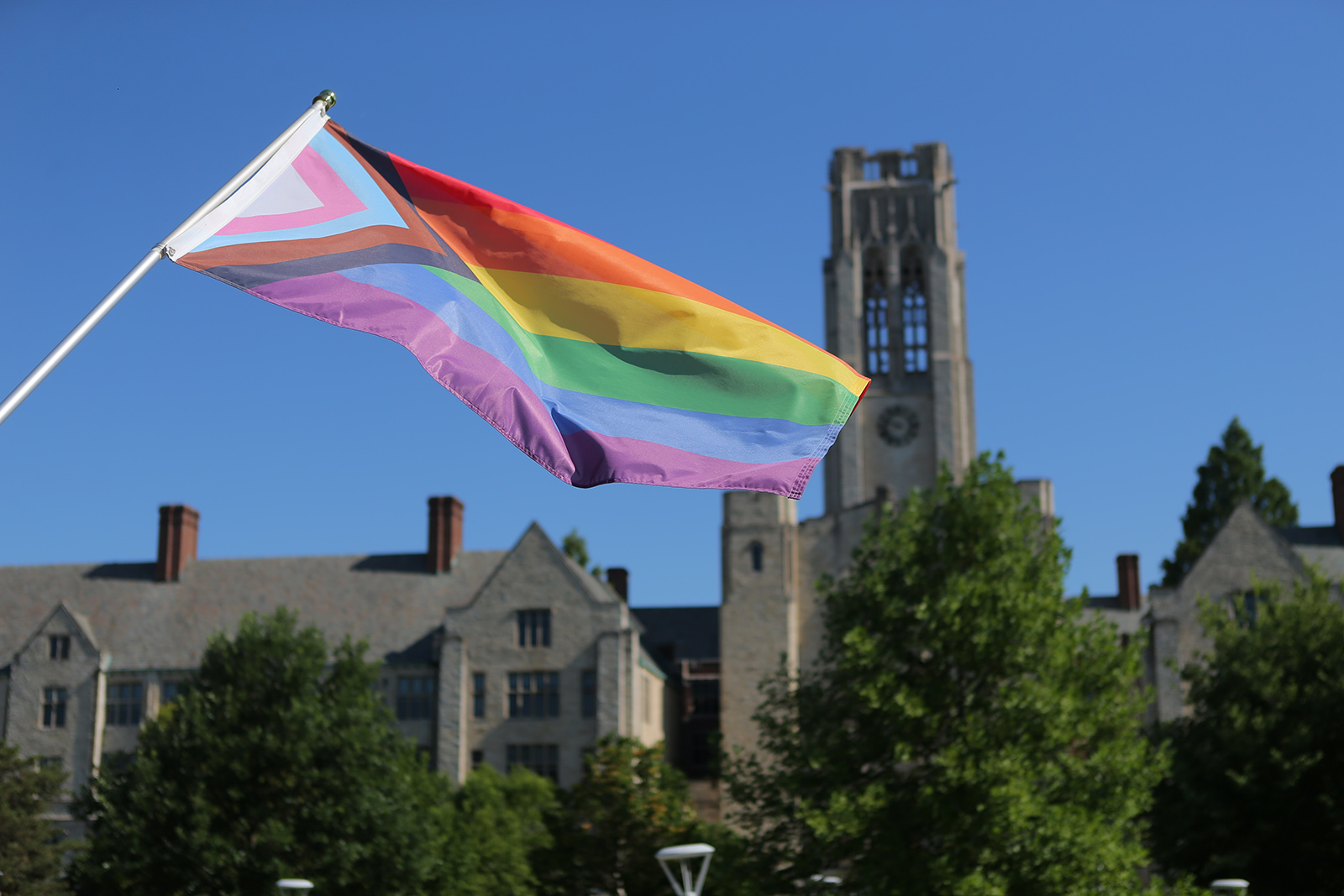 A photo of a Pride Progress Flag being waved with University Hall on Main Campus in the background.