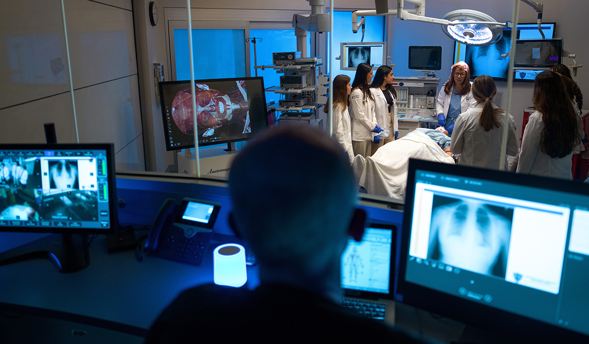 Instructor leads a group of medical students in white coats around a simulated patient in a high-tech operating room at The University of Toledo’s Interprofessional Immersive Simulation Center, as a technician monitors the session from a nearby control room with multiple screens showing medical imaging.