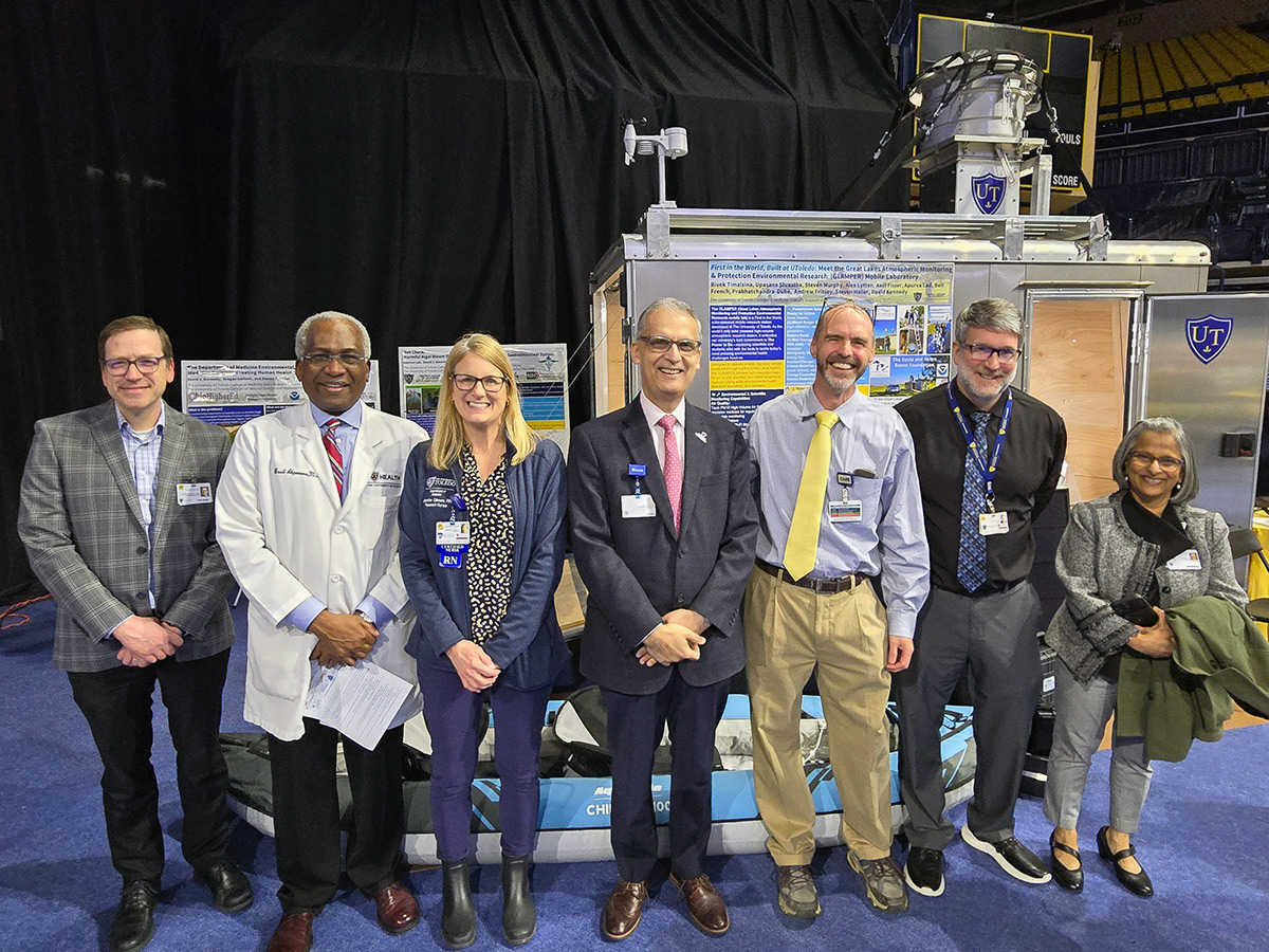 Leaders from the College of Medicine and Life Sciences pose for a group photo with researchers Drs. Kennedy and Haller in front of their Great Lakes Atmospheric Monitoring & Protection Environmental Research (GLAMPER) Mobile Laboratory at the R1 research celebration in Savage Arena. 