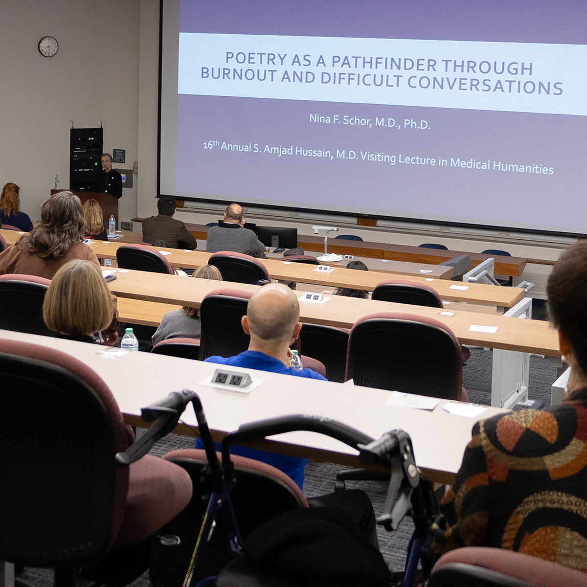 Dr. Nina F. Schor stands at a podium with attendees in the classroom at the beginning of the lecture. On the screen is her title slide showing the title of her speech, "Poetry as a Pathfinder through Burnout and Difficult Conversations."