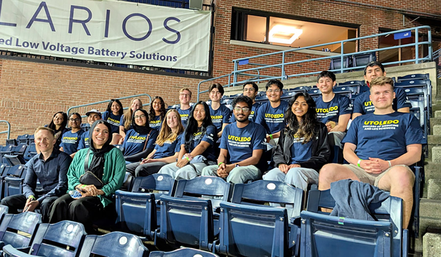 A group of MedStart students and program leaders pose in stadium seats at a Toledo Mud Hens game, wearing matching navy UToledo College of Medicine and Life Sciences shirts.