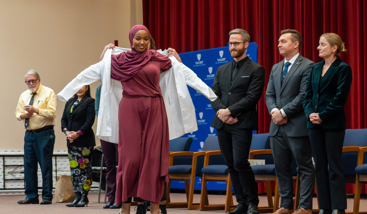 Graduating Physician Assistant student receives a white coat during the End of Program Ceremony, with faculty and program leaders standing behind them on stage.