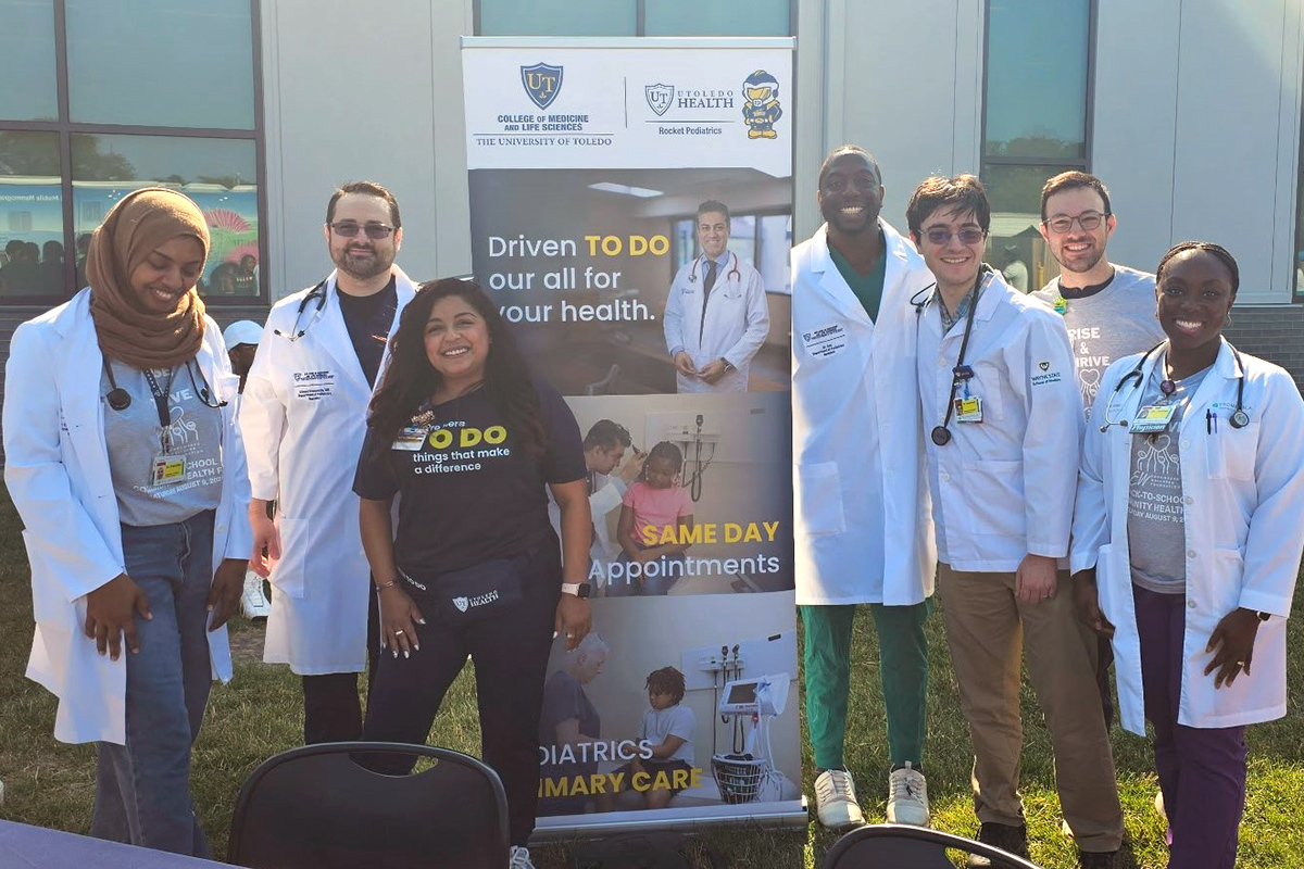 Seven members of the UToledo Department of Pediatrics pose for a group photo around the UToledo branded sign during the Rise and Thrive Community Health Fair at the Wayman Palmer YMCA. The sign promotes UToledo Health and the College, with text: Driven To Do our all for your health; same day appointments; pediatrics primary care. 