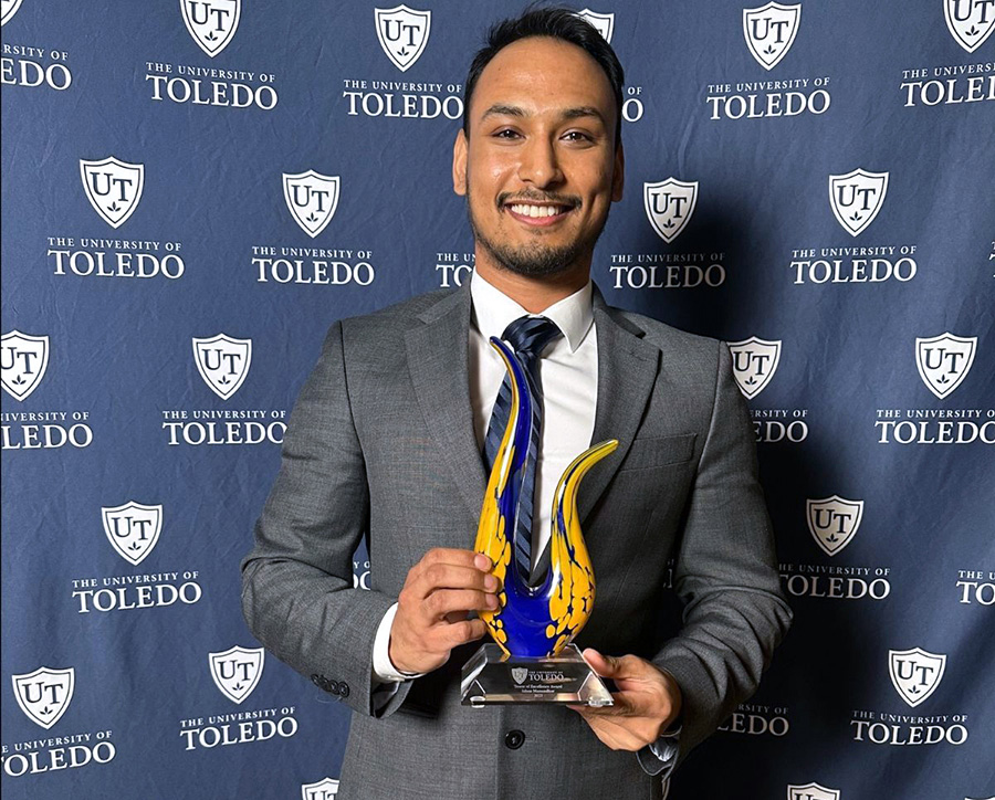 Graduating student Ishan Manandhar holds a colorful glass trophy for the Tower of Excellence Award and stands in front of a blue UToledo background. 