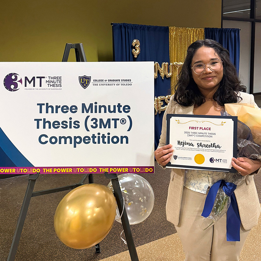 Rejina Shrestha stands smiling and holding a first-place certificate and a bouquet in front of a Three Minute Thesis (3MT) competition sign and decorative backdrop.