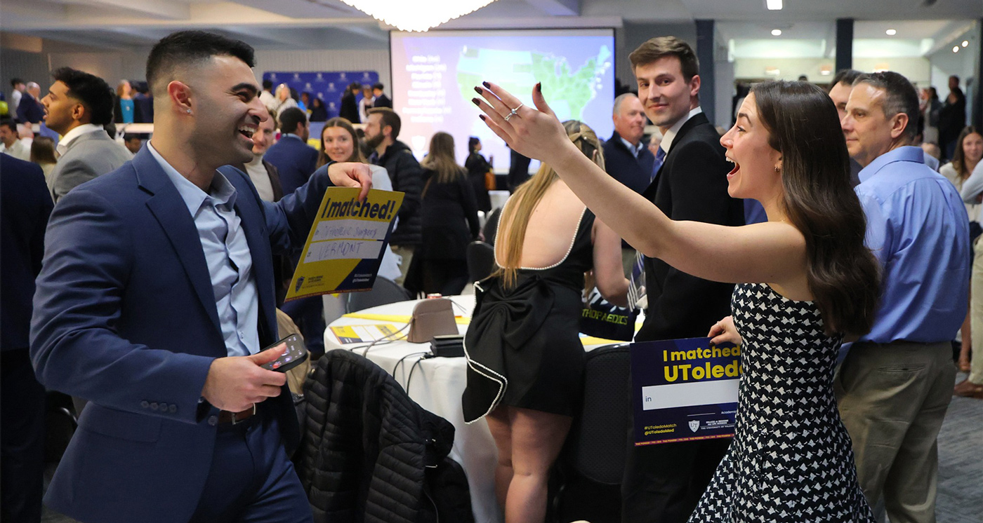 Two medical students celebrating their Match Day results in a crowded event space, smiling and gesturing excitedly while holding a yellow and blue "I matched!" sign.