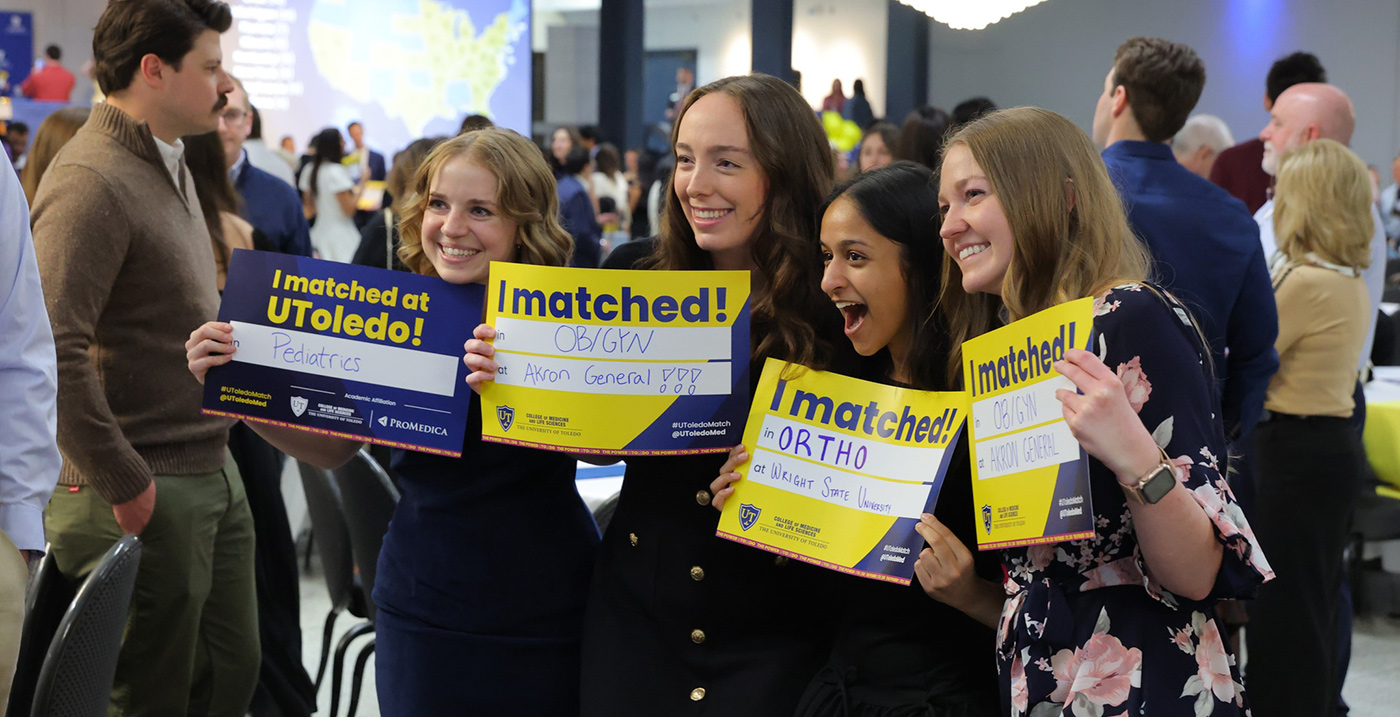 Four medical students smiling and posing together at a Match Day celebration, each holding a yellow and blue "I matched!" sign with their specialty and residency location written on it