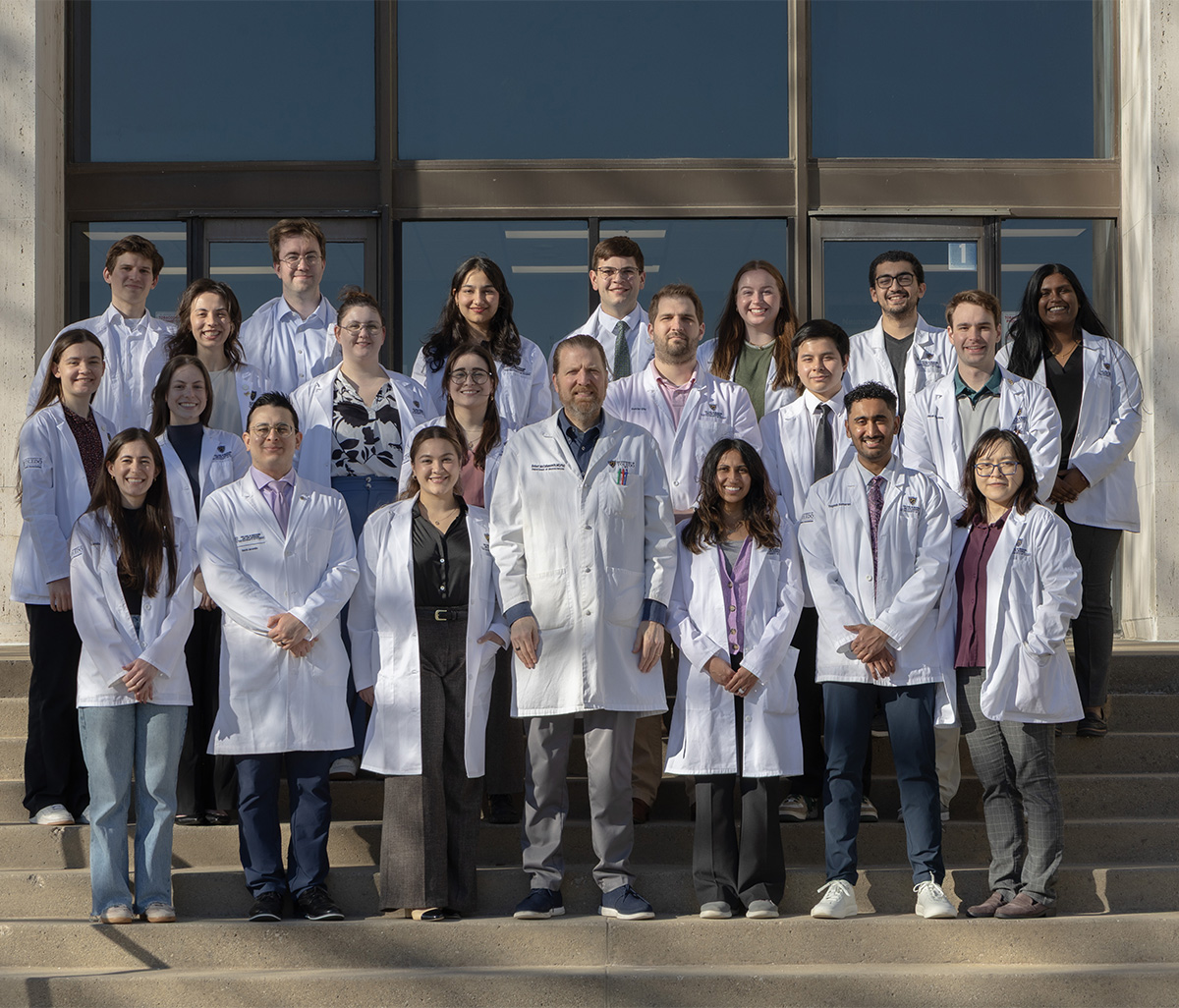 A group photo of M.D./Ph.D. students on steps in front of Block Health Science Building in white coats.