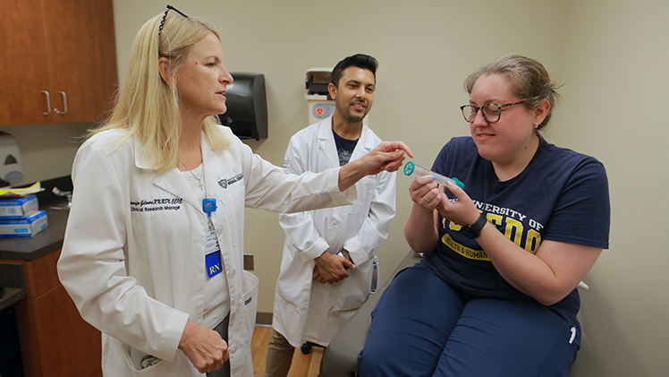 Three people are inside a clinic room. Jennifer Gilmore, left, demonstrates one of the lung screening devices that will be used in the Great Lakes Aerosol Monitoring Research Study as Katie Behrens plays the role of a patient.
