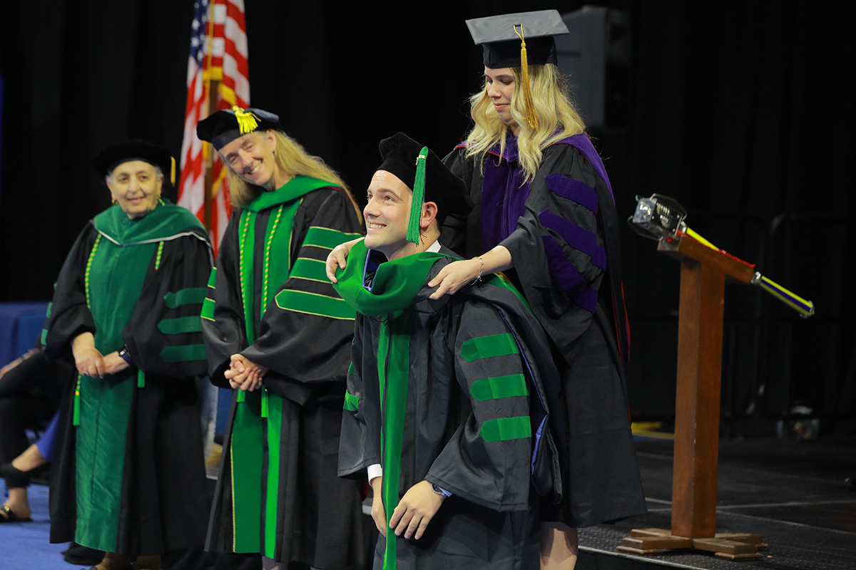 A graduate is hooded with faculty looking on in the background and smiling.  