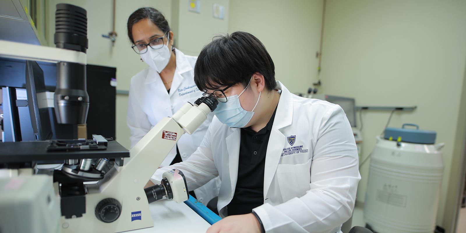 Student looks into microscope while being supervised by a faculty member.