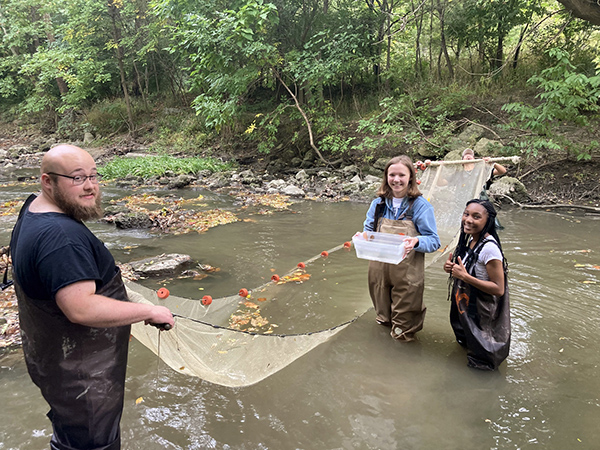Three students standing in a river wearing water proof gear. The students are cleaning up the riving using nets and bucks to collect material from the water