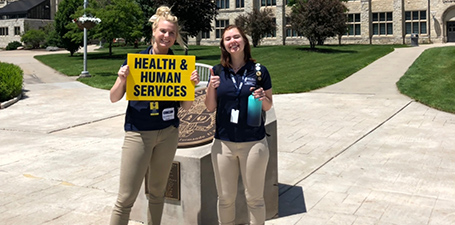 Two students standing the the mall with sign for Health and Humas Services