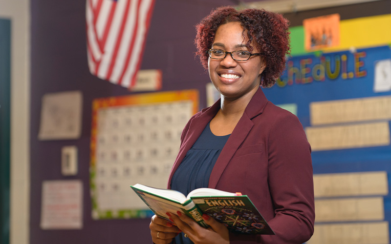 smiling teacher stands in classroom holding an English textbook