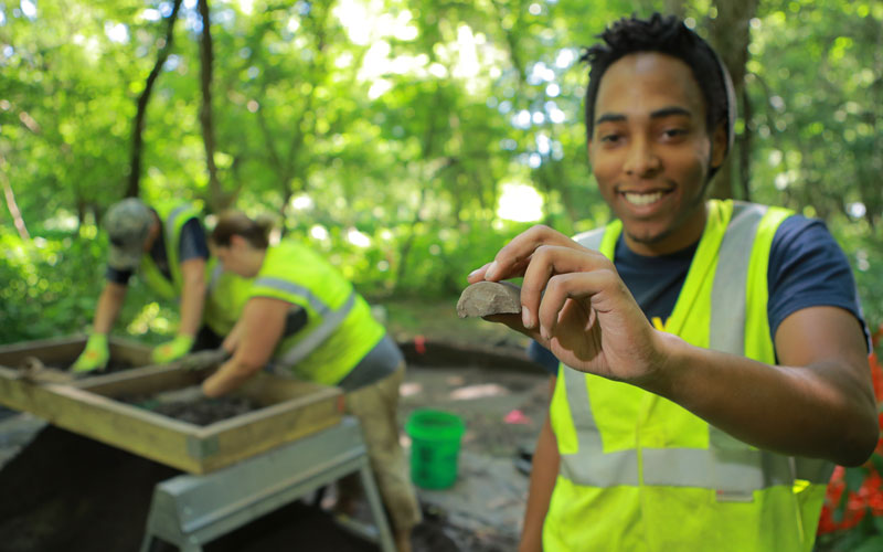 student wearing a neon vest at a dig site holds up and observes a rock while two students in the background sift through a pile 