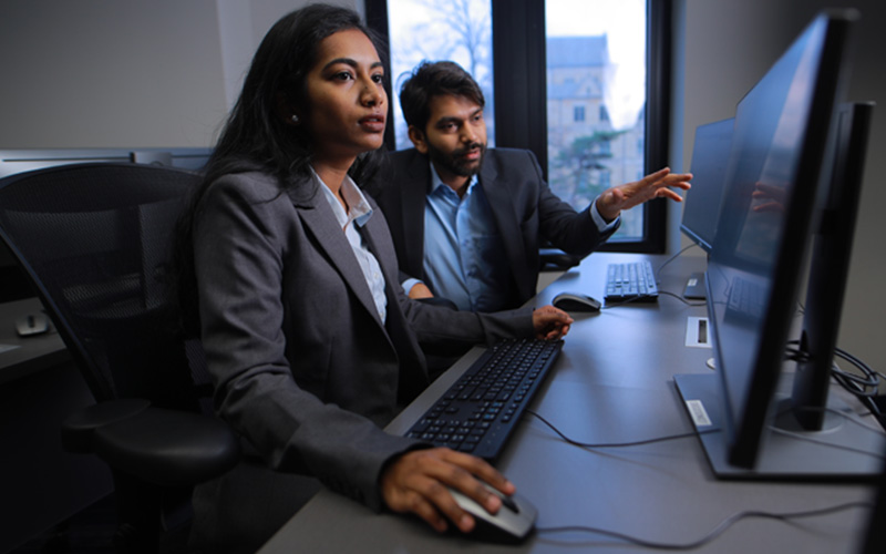 a woman in a computer labs observes her screen the man sitting besides her points to the computer monitor