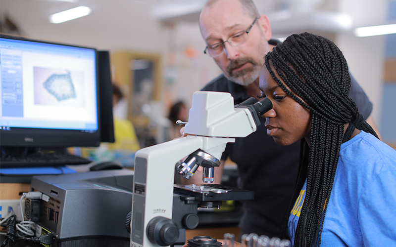 Woman in a lab looking though a mircoscope while the professor adjusts the microscope
